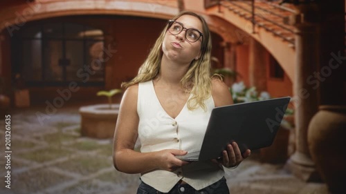 Woman holding laptop with pursed lips and puffed cheeks in a terracotta courtyard; puzzlement curiosity.