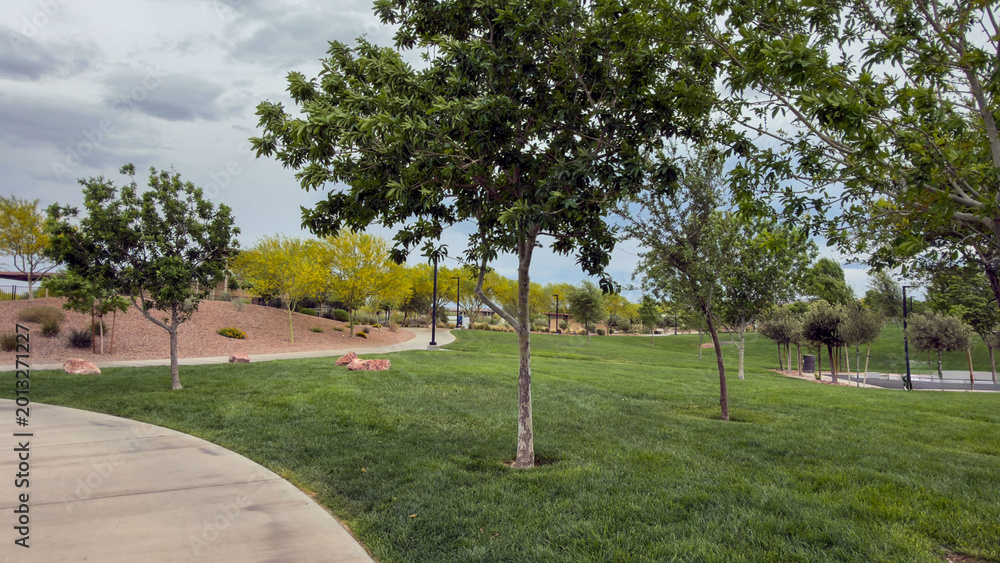 Obraz premium Vibrant view of lush green hills and manicured lawns in a public park. Featuring winding walking paths, decorative shrubs, and leafy trees under a clear blue sky. Peaceful summer day atmosphere.