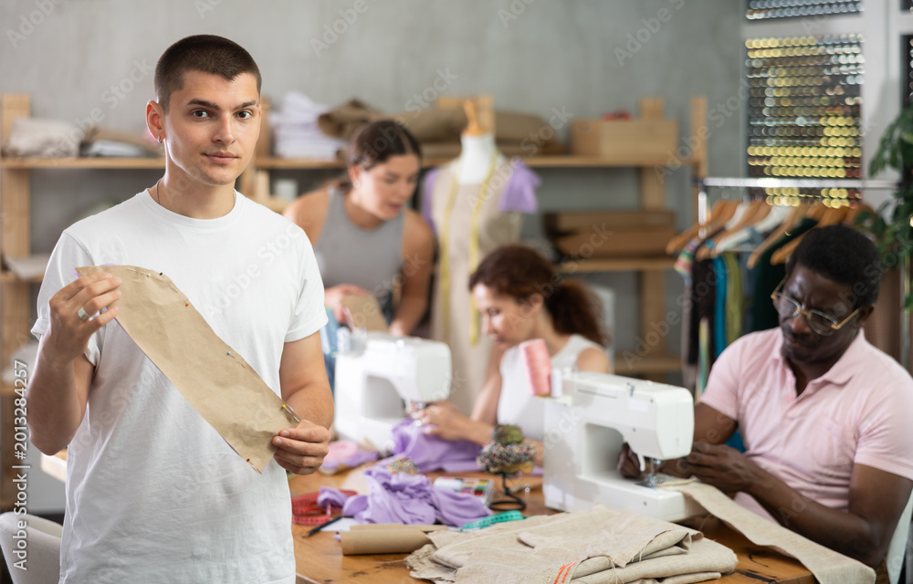 Obraz premium In training tailoring workshop for adult beginners, engaged young guy reviewing paper pattern pinned to fabric surrounded by enthusiastic classmates working on different sewing tasks