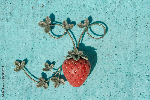 A decorative strawberry ornament with detailed leaves casts a crisp shadow on a bright turquoise texture background, creating a vintage, whimsical still-life composition with bold color contrast.