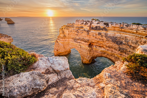 Sunrise view of Praia da Marinha beach in Algarve, Portugal, with a limestone sea arch, Atlantic water, warm sky, and rugged coastal cliffs.