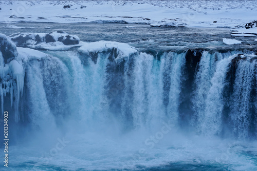 Schilderij op canvas Icy Waterfall in Winter Landscape With Snow Covered Cliffs and Misty Blue Waterf