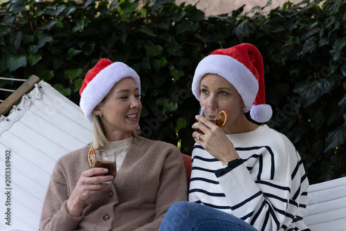 Billede på lærred Female friends sitting on white hammock bench in backyard wearing Santa hats hol
