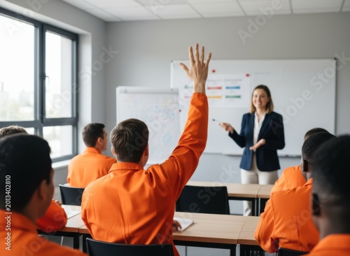 A group of individuals in orange uniforms in a classroom setting, with one person raising their hand to a female instructor. They are seated at desks, engaged in a learning environment.