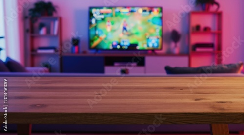 Close up wood table surface in modern room. Blurred background shows TV playing game with neon pink purple light. Empty room for product display.