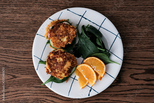 A stylish breakfast plate with two carrot cakes on fresh spinach leaves, accompanied by cucumber slices and vibrant orange wedges, served on a white plate with blue grid lines on a wooden table.