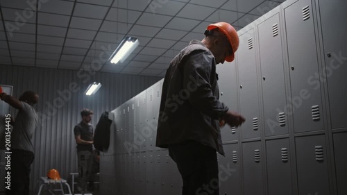 Male Professional in Safety Helmet and Workwear Enters Locker Room, Friendly Greets Coworkers, and Starts Changing After Long Shift in Modern Factory. Concept of Professional Industrial Routine.