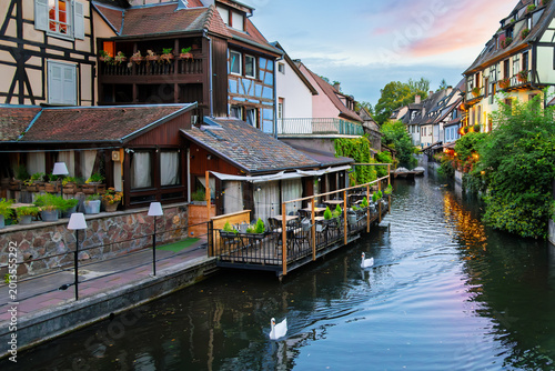 Принти на полотні Swans float past half timbered medieval buildings with waterfront cafes along the narrow Lauch River canal in the Petite Venise canal district of Colmar, France