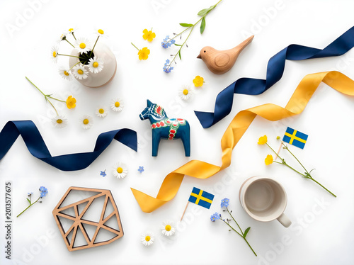A minimalist flat lay featuring blue and yellow Swedish elements, delicate white daisies, flowing silk ribbons in navy and gold, and iconic Scandinavian design objects like a small dala horse