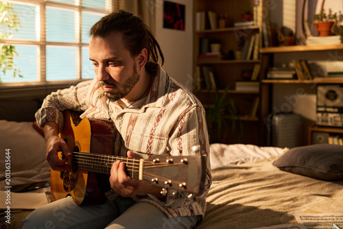 Bild auf Leinwand Young adult Caucasian man playing acoustic guitar while sitting on bed, practicing songwriting at home and expressing creativity