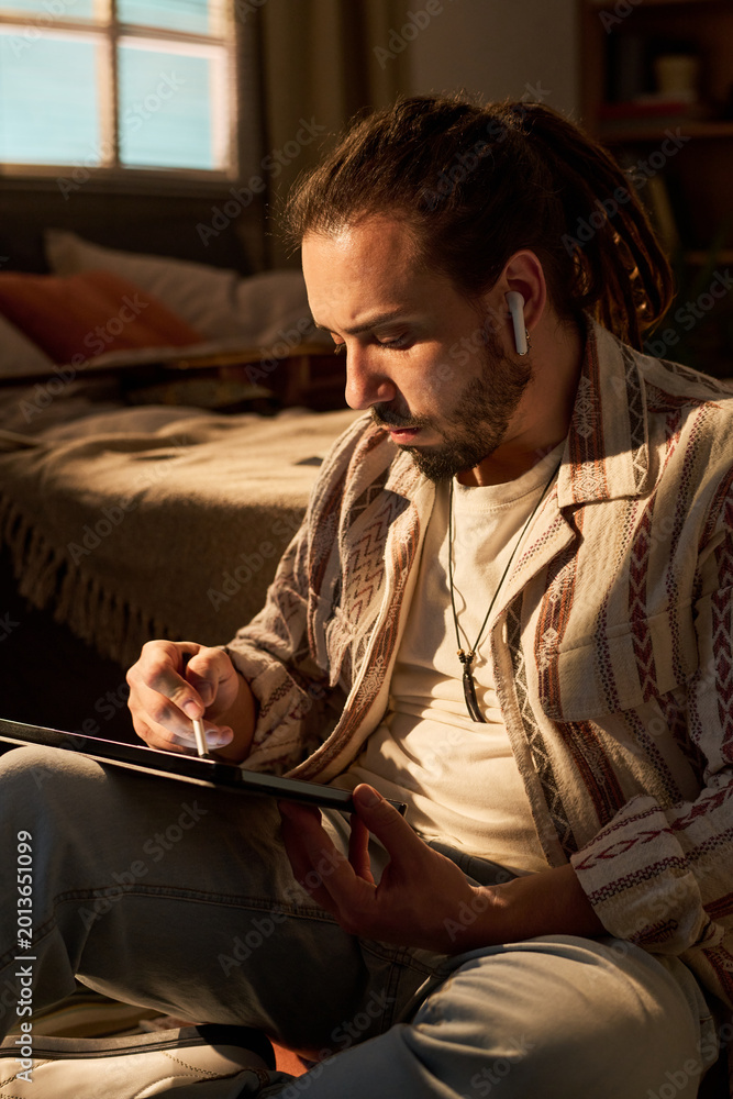 Obraz premium Vertical shot of young adult man using tablet while wearing wireless earbuds at home, browsing digital content. Suitable for remote lifestyle, streaming service, or mobile app marketing