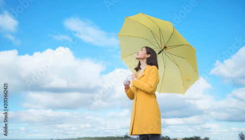 Woman in yellow coat holds matching umbrella looking up at bright blue sky with soft clouds, feeling peaceful and hopeful