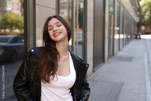 A beautiful brunette girl in a leather jacket, jeans and sneakers on the street of a modern city.