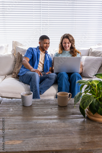 Diverse couple sitting on light-colored sofa sharing silver laptop on lap near wooden coffee table