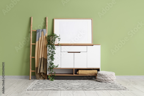 Foto Interior of living room with blank frame, white commode and plaid on ladder near