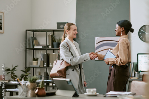 Medium shot of Caucasian businesswoman greeting Black businesswoman with handshake during office meeting, signaling partnership and hiring. Ideal for recruitment, onboarding, dealmaking