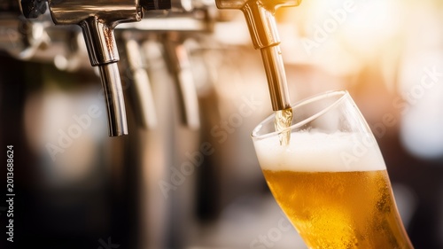 Close up of refreshing golden lager beer is poured from a professional metal tap into a clear glass creating thick white foam during a busy evening, night at pub, bar indoors
