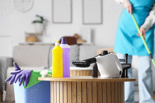 Принти на полотні Steam cleaner with supplies on table against female janitor mopping in room