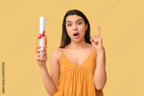 Surprised female student with raised index finger holding diploma on yellow b...