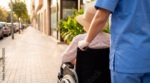 Cuadro en lienzo Healthcare professional in gloves assists a senior woman in a wheelchair on a sunny street, providing mobility