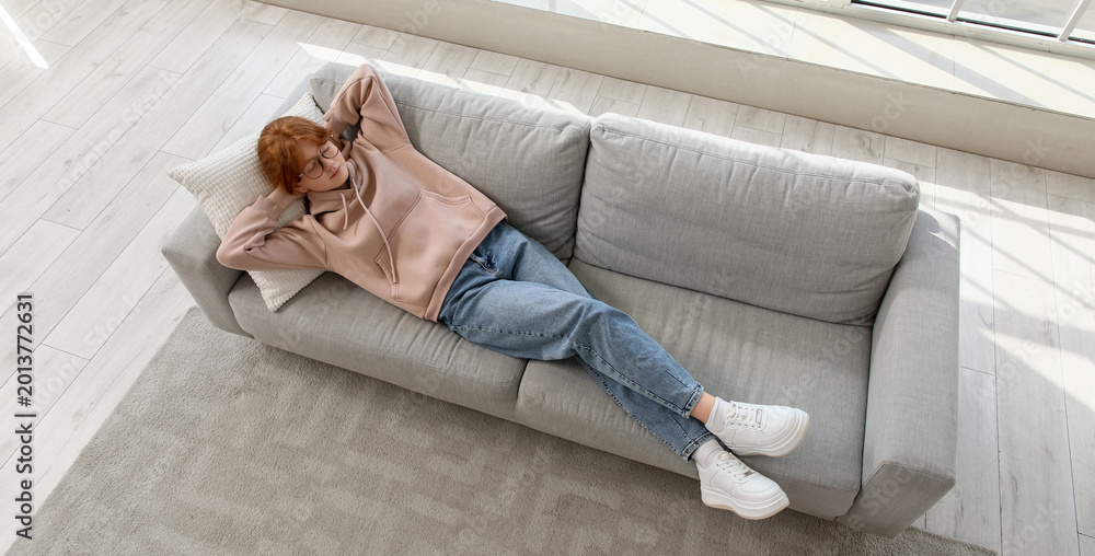 Fototapeta premium Redhead teenage girl lying on sofa near large window at home, top view
