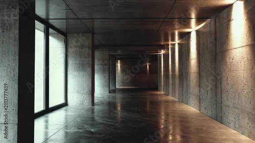 Long and empty modern interior corridor featuring raw concrete walls, large windows letting in natural light, and illuminated recessed lights casting reflections on the wet, dark floor