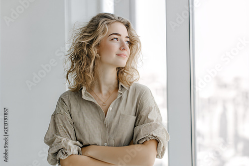 Caucasian freelance designer with curly blonde hair and freckles, arms crossed in striped shirt with rolled sleeves, gazing out sunlit studio window