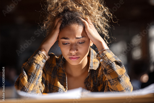 African American college student in yellow plaid shirt clutching her temples over scattered study papers, curly ponytail, anxious and fatigued