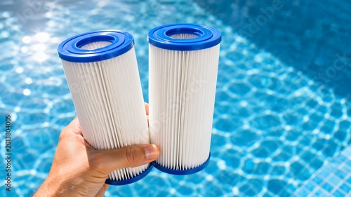Hand holding two pleated swimming pool filter cartridges over blue water surface, pool maintenance equipment with water filtration system replacement in summer outdoor