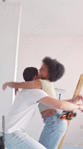In a sunlit room, a couple embraces while working on a home renovation project, she's wearing a tool belt, he's casually dressed in a white t-shirt and jeans.