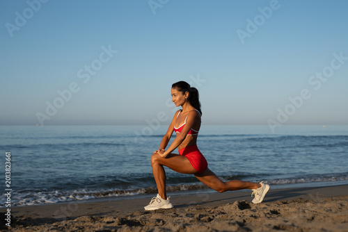 Focused athletic woman in red sportswear doing deep lunge stretch on sandy beach at sunrise, facing sideways toward ocean, enjoying peaceful morning workout by the sea