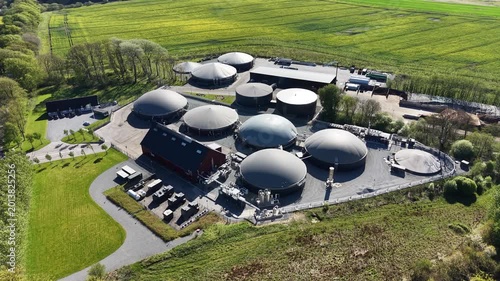 Close-up drone shot of circular anaerobic digesters at a biogas facility surrounded by yellow blooming rapeseed fields under bright sunlight.