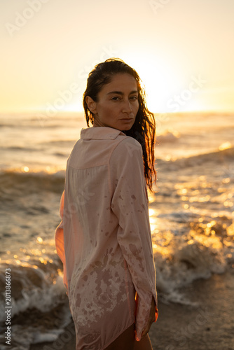 Portrait of a woman standing in sea waves at golden sunset, wearing a wet shirt, backlit by warm sunlight, creating a serene and cinematic beach mood