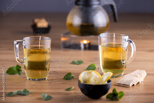 Fresh green tea served in transparent glass cups with lemon slices, mint leaves and glass teapot on wooden table. Healthy herbal beverage concept with elegant studio lighting and cozy atmosphere.