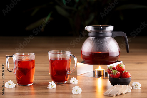 Warm red fruit herbal tea served in transparent glass cups with strawberries, tea bags and glass teapot on wooden table. Cozy beverage concept with soft candlelight and elegant studio lighting.