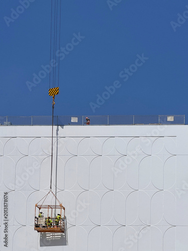 construction site with men standing inside cage on a crane against white wall on new building exterior