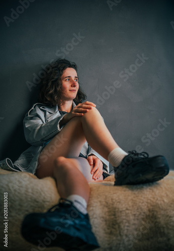 Playful young girl with beautiful legs seated against a textured wall in a denim jacket and shorts, captured in soft natural lighting, radiating a relaxed and introspective vibe of youth.