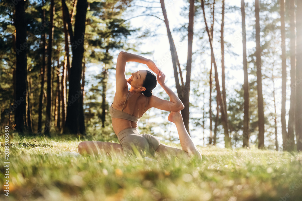 Fototapeta premium Female yoga practitioner performing a backbend pose on grass in a forest setting with tall trees and sunlight filtering through the branches