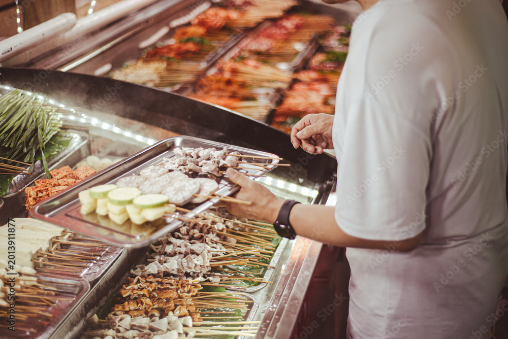 Obraz premium Chilled skewer display features lotus root, zucchini, tofu, and assorted meats arranged in refrigerated trays while a shopper builds a tray of chosen items, Mengzi, Yunnan