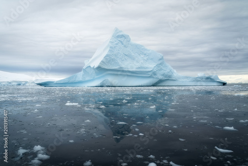 View of a massive pointed iceberg floating in the calm dark waters with smaller ice fragments and a reflection under an overcast sky in Antarctica.