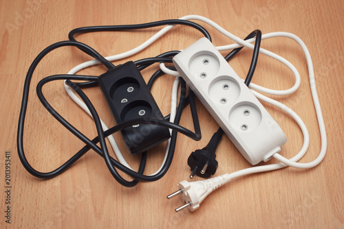 Tangled black and white household power extension cords with multiple electrical outlets on a floor. The concept of electricity safe usage.