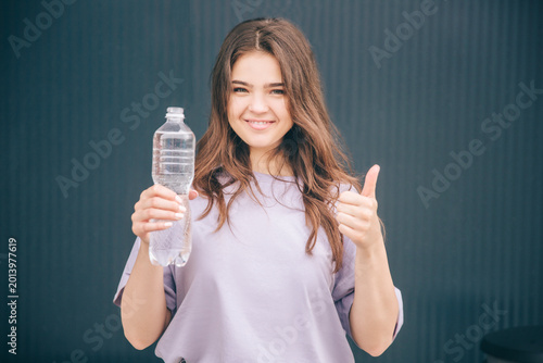 Young stylish trendy woman isolated over grey blue background. Cheerful girl holding plastic water bottle and showing big thumb up. Clear good water. Healthy and take care of health.