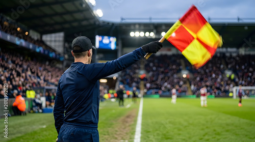 Assistant referee holding a flag on the sideline during a professional football match in a stadium with spectators.