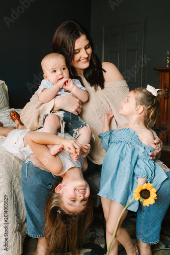 Smiling mother with three young children playing joyfully on bed, one child upside down, cozy family moment