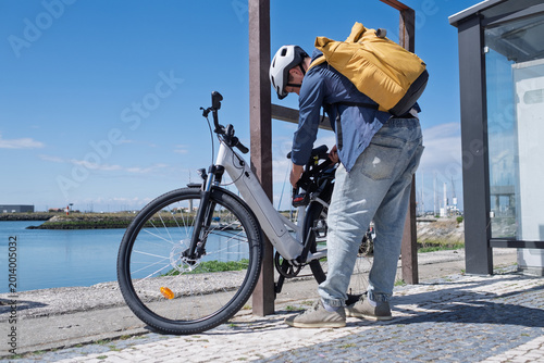 Young man in helmet securing his electric bicycle with a lock at a city bike rack near the water