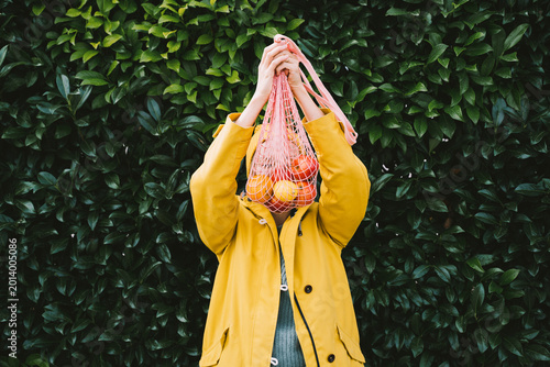 Cuadro en lienzo Young woman wearing yellow hat and raincoat holding bag with fresh organic vegetables against green leaves wall
