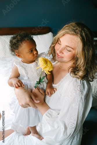 Loving mother holding young daughter in white dress with yellow flower, tender moment indoors

