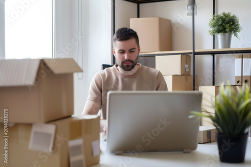 A focused male small business owner sitting at his desk using a laptop, surrounded by stacked cardboard boxes ready for shipment in a bright home office or warehouse setting