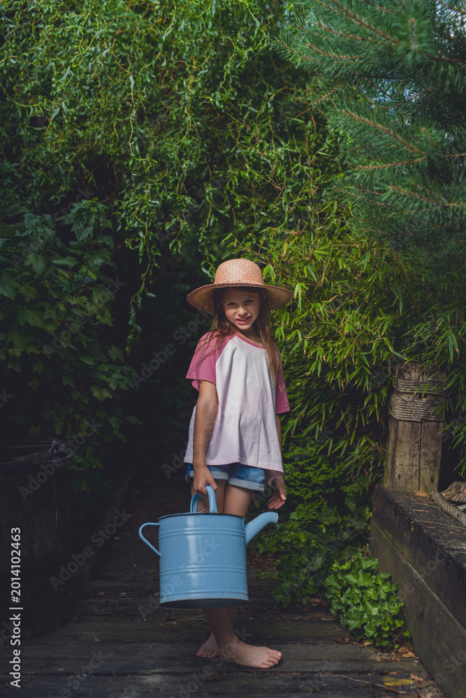 Fototapeta premium Little girl holding watering can in lush garden pathway.