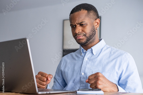 Foto A frustrated man in his home office clenches his fists while staring at a laptop, expressing stress and annoyance during remote work, technical difficulties, or a tense online meeting